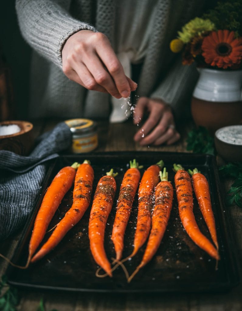 Side view of person sprinkling salt onto a tray of whole carrots on a sheet pan