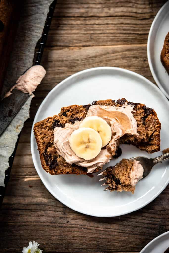 Overhead view of slice of banana bread on white plate with cinnamon cream cheese and banana slices