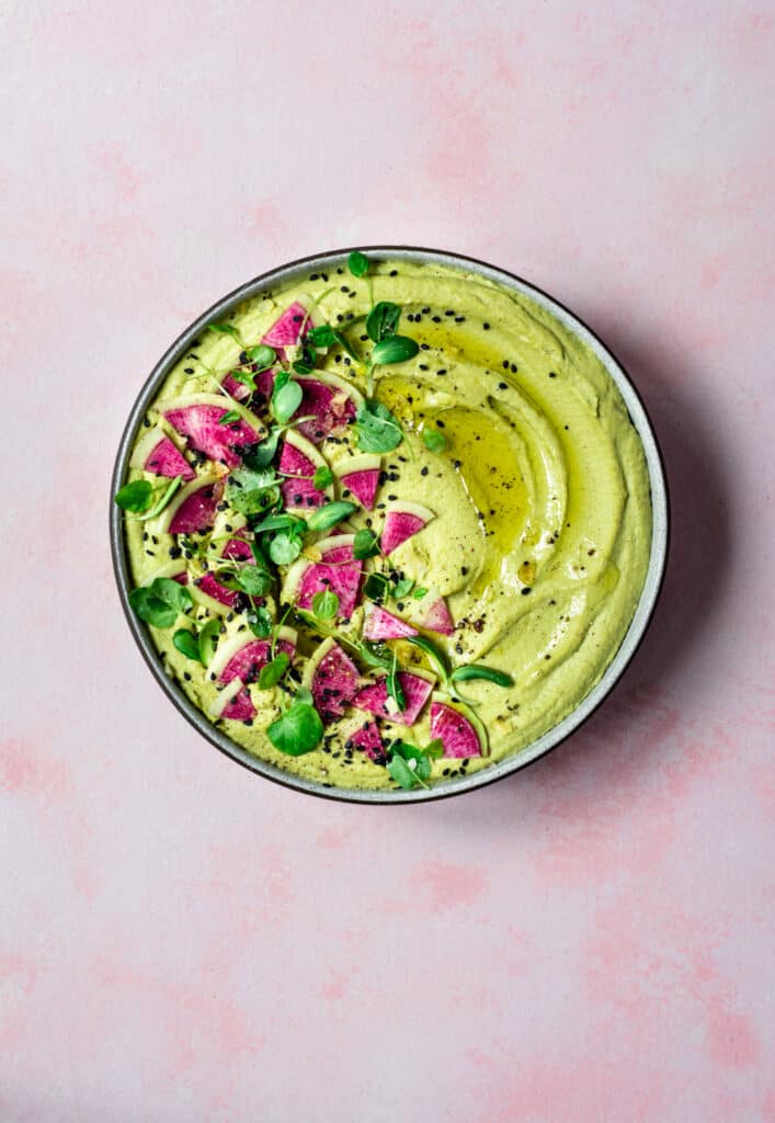 Overhead of green romanesco hummus topped with watermelon radish in a white bowl on pink background