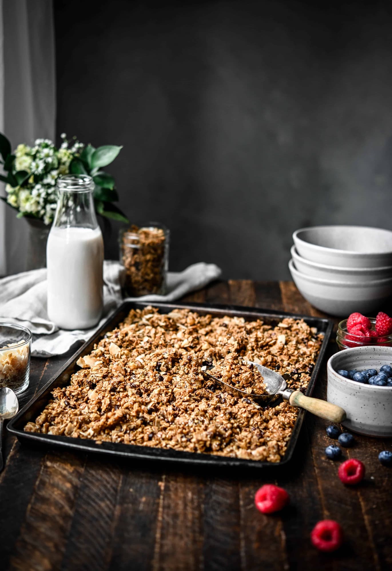 Side view of a baking sheet with granola and a scoop on a wood table with greenery in background