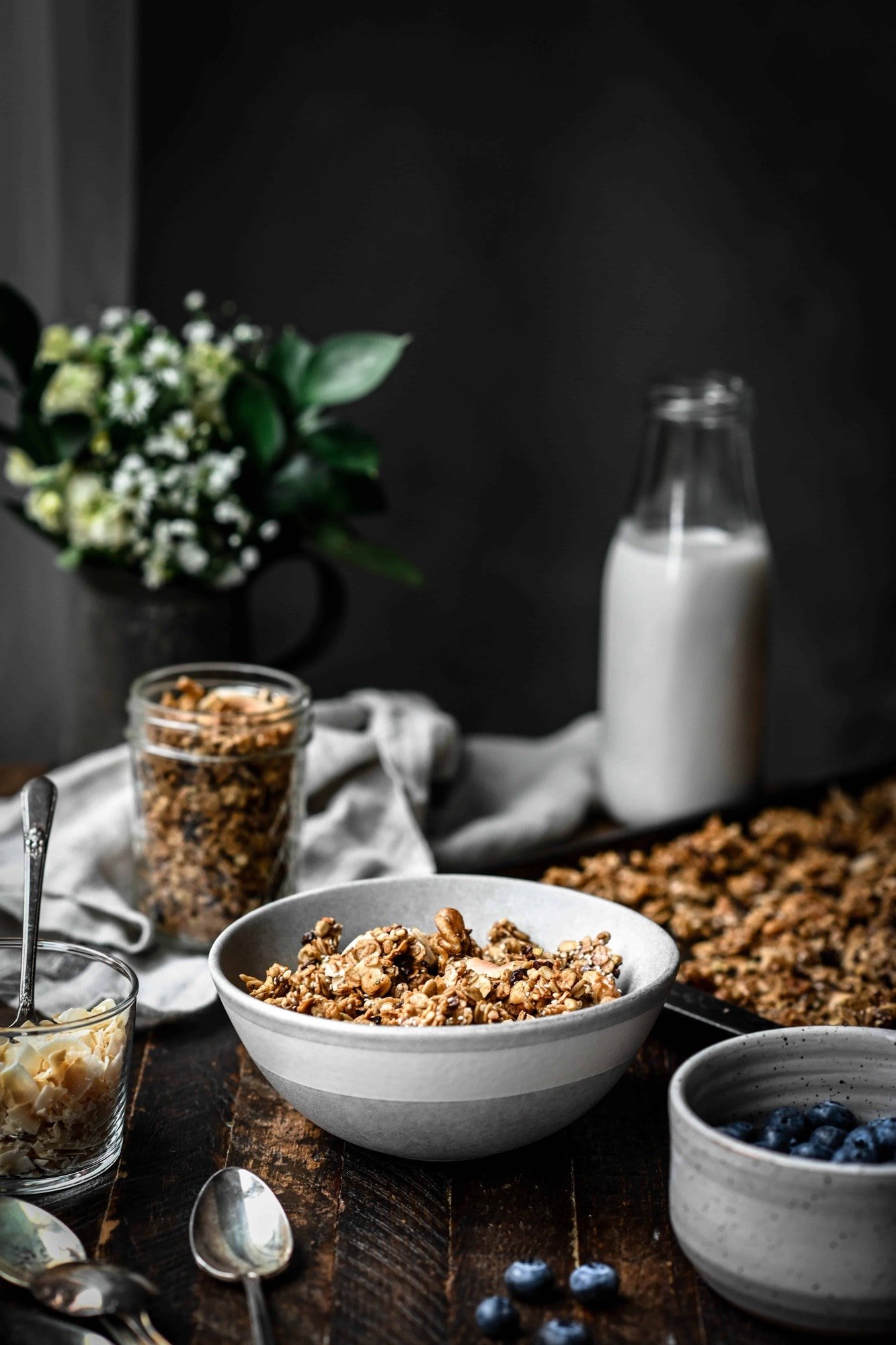 Side view of a bowl of gluten free salted caramel granola on a wood table with more granola, milk and flowers in background