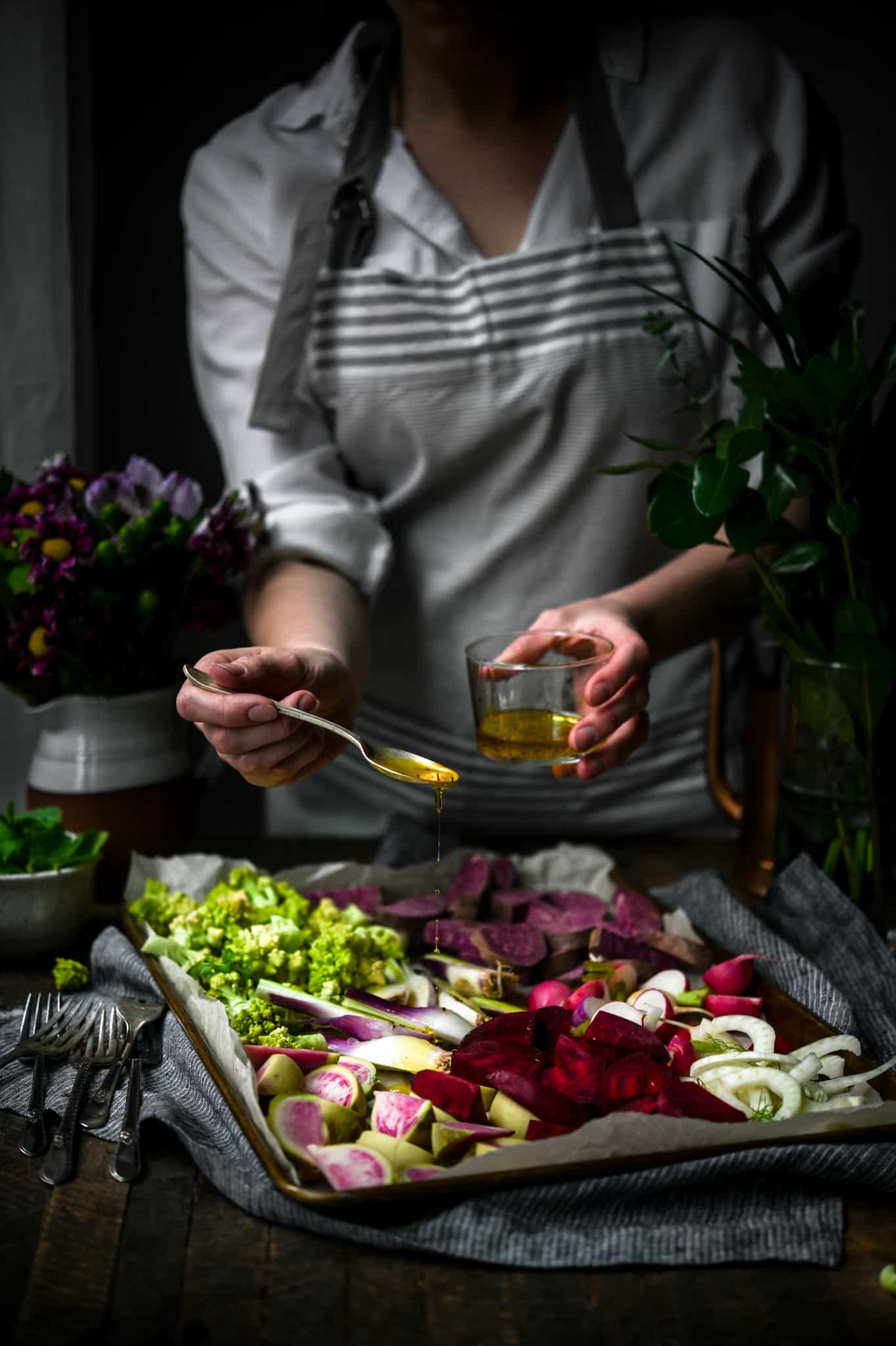 Person in apron drizzling olive oil onto sheet pan filled with vegetables