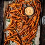 Overhead of spiced sweet potato fries on an antique baking sheet with fresh herbs and aioli