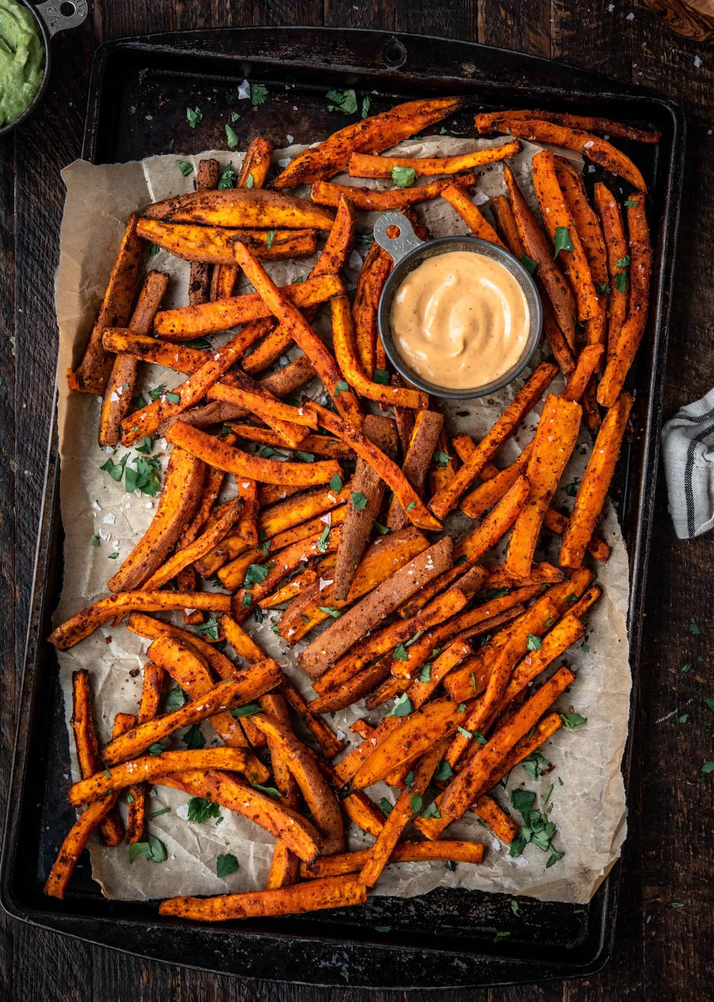 Overhead of spiced sweet potato fries on an antique baking sheet with fresh herbs and aioli