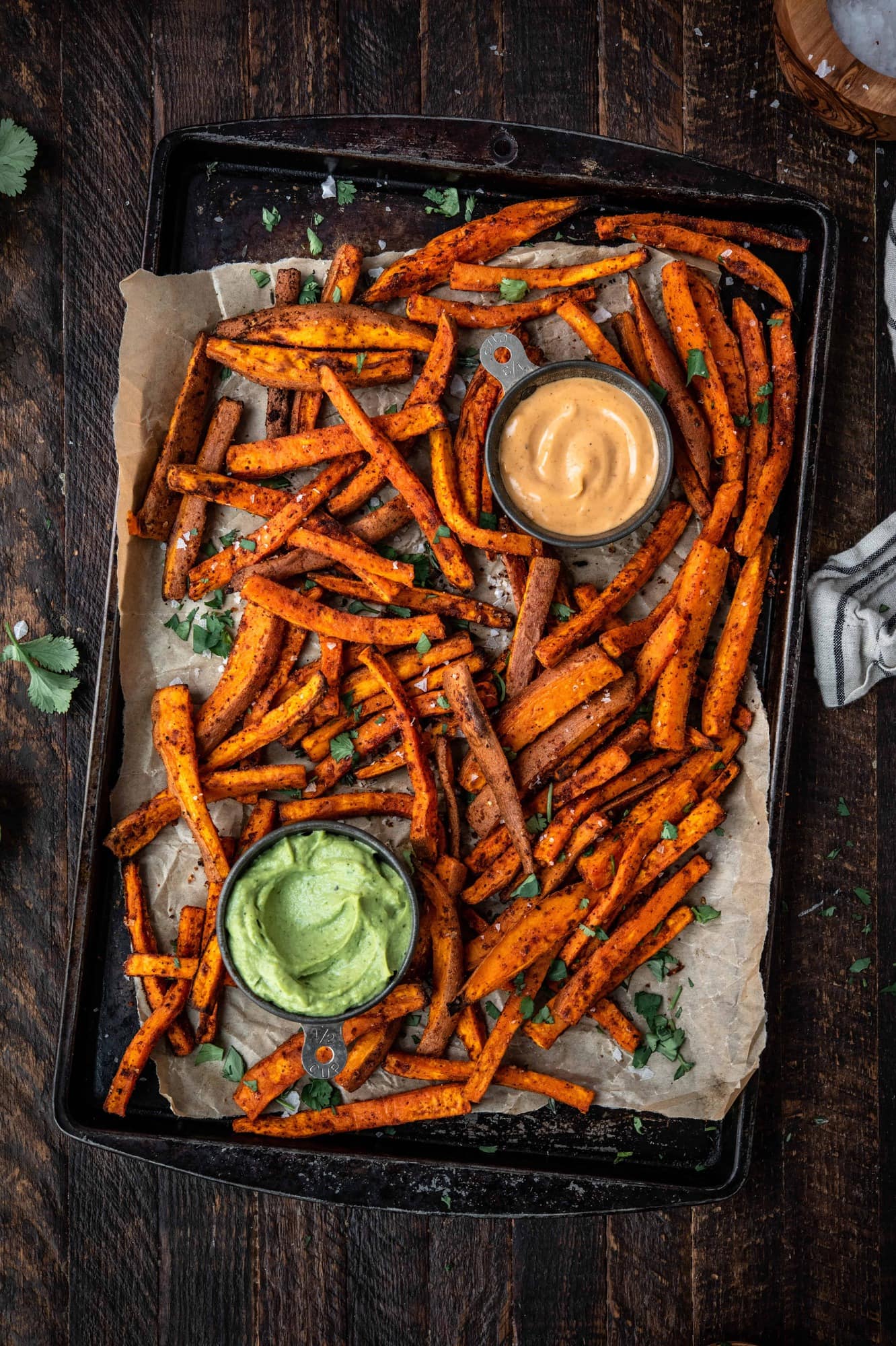 Overhead of spiced sweet potato fries on an antique baking sheet with fresh herbs and aioli