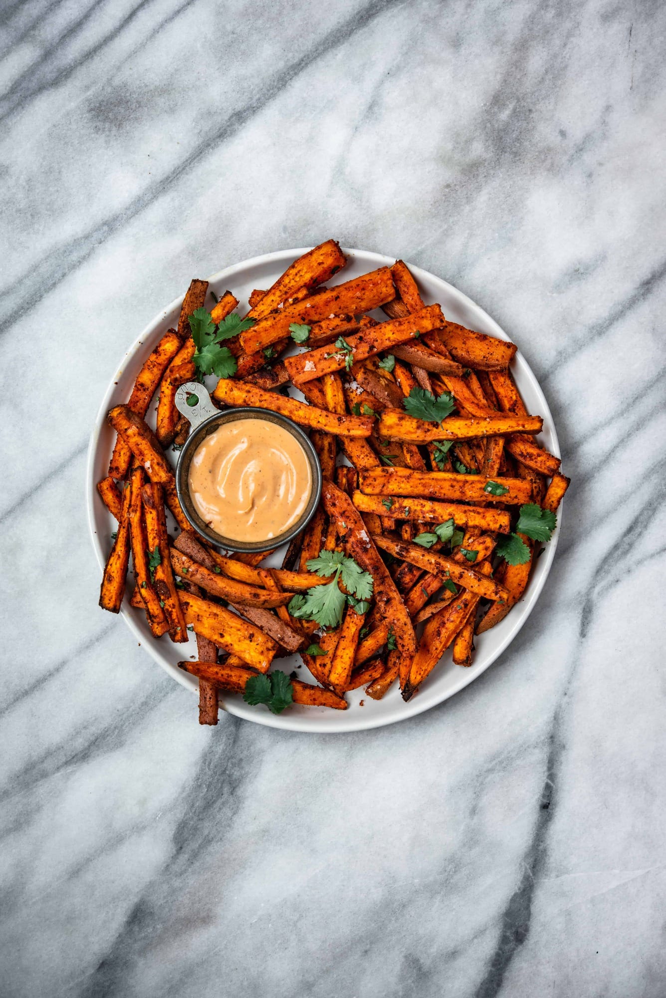 Overhead of spiced sweet potato fries on a white plate with fresh herbs and aioli on a marble background