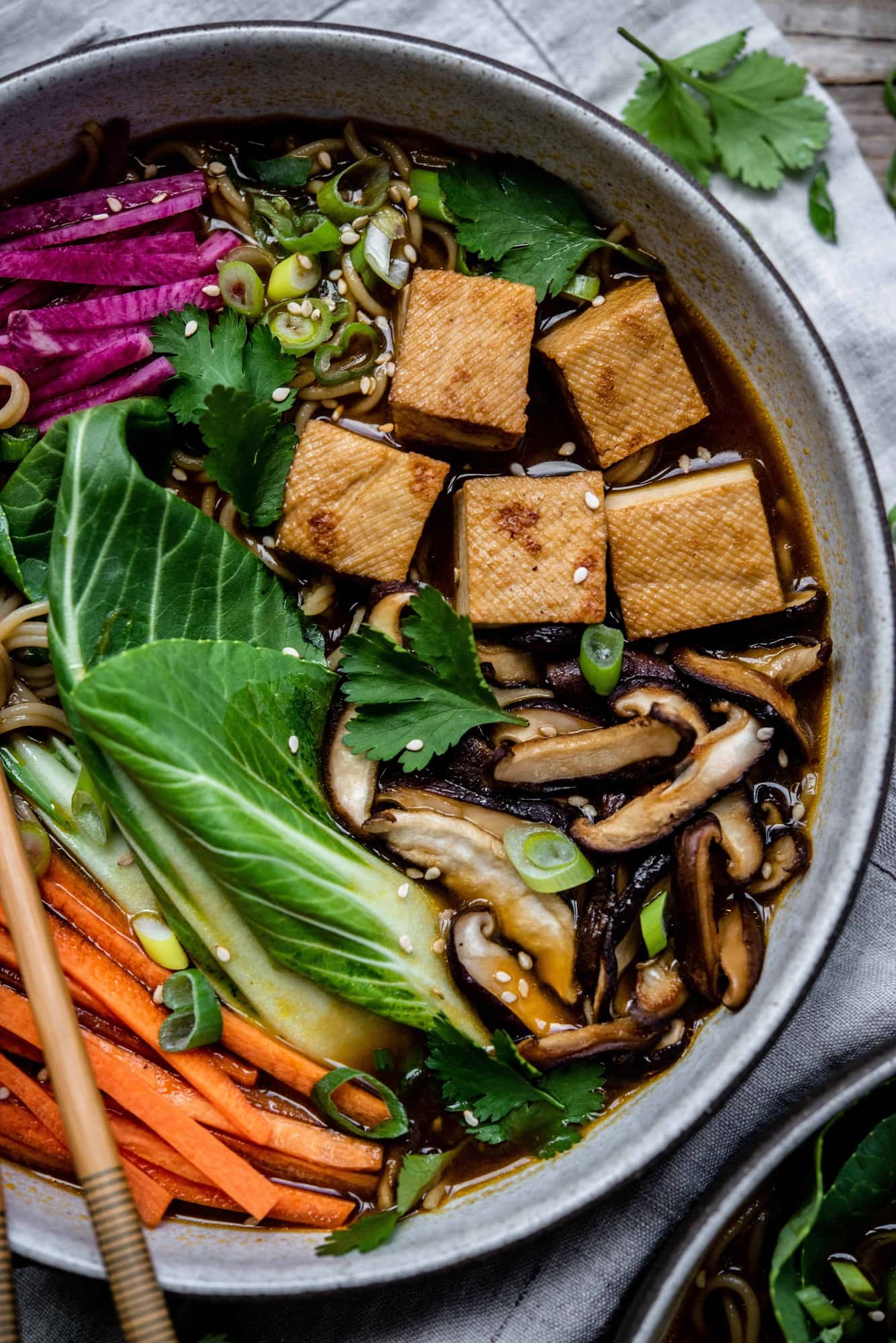 Close up of homemade vegan ramen in a white bowl with chopsticks, bok choy, tofu, mushrooms, carrots and noodles