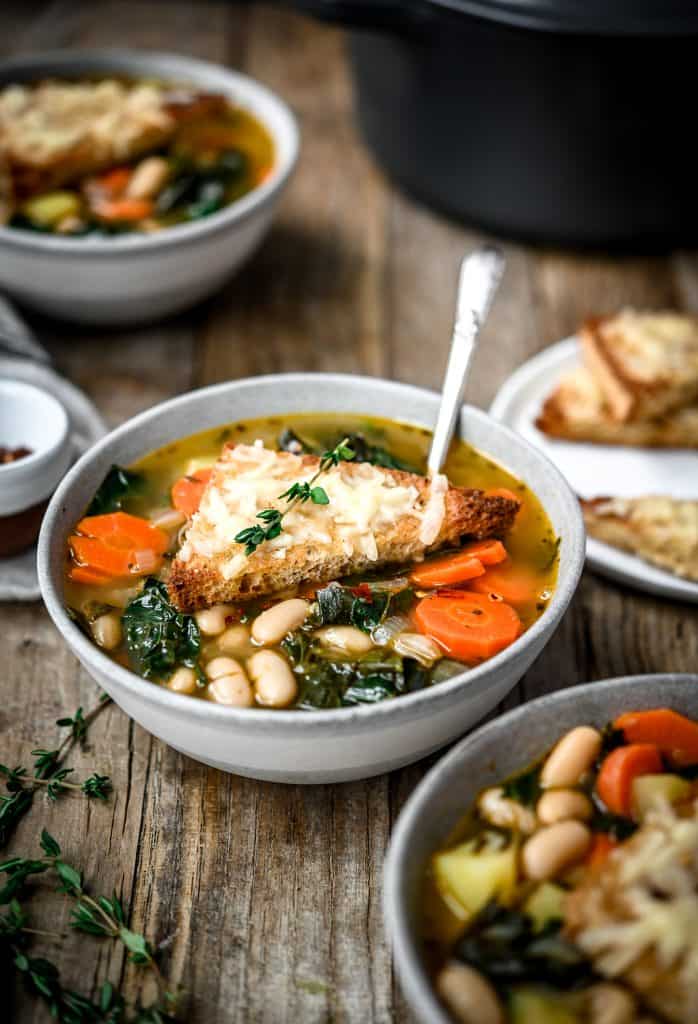 Side view of one-pot white bean kale soup in a grey bowl on a wood table
