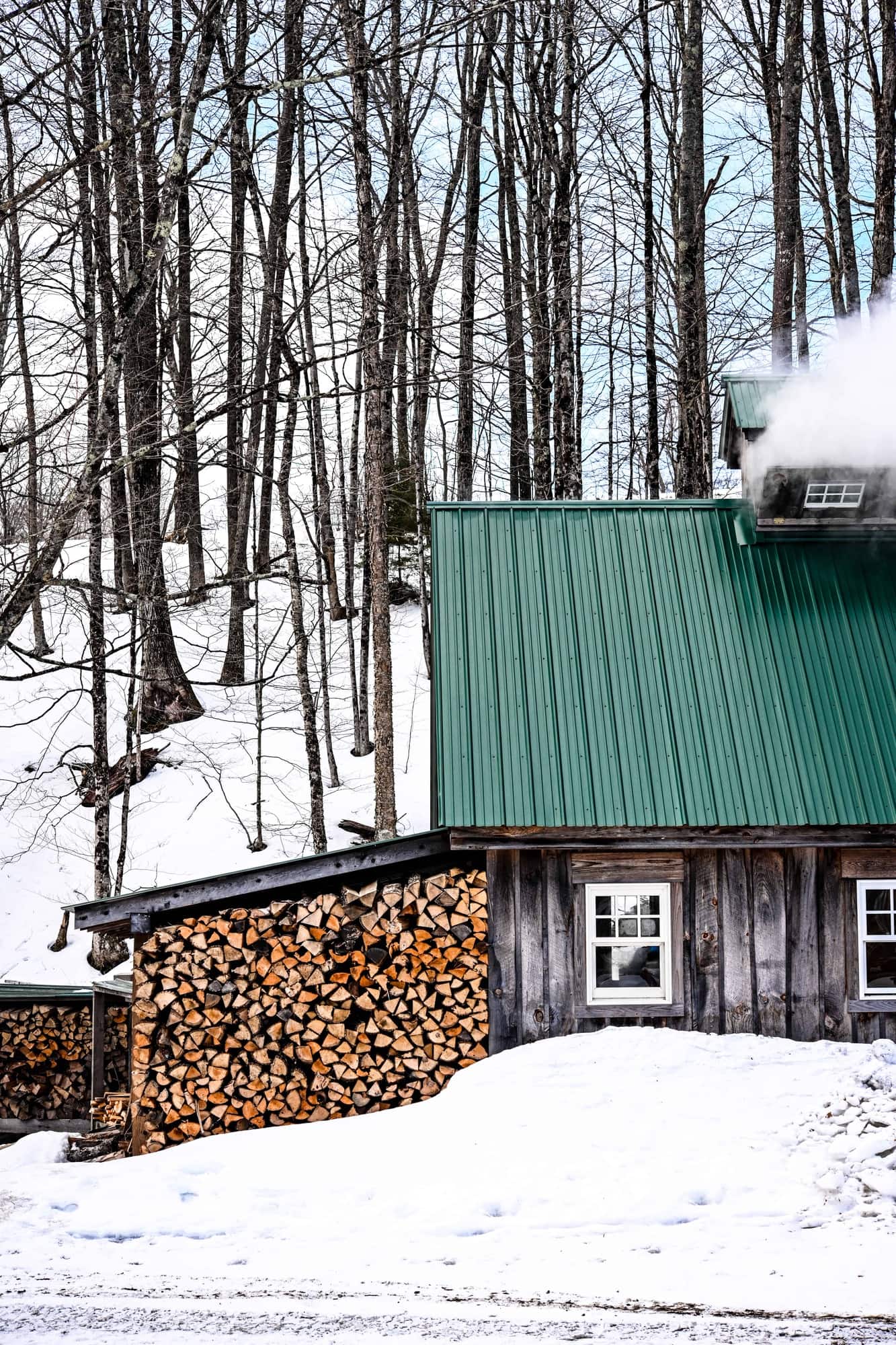 Side view of a Vermont Sugarhouse in winter with stack of wood and steam coming out of top of house