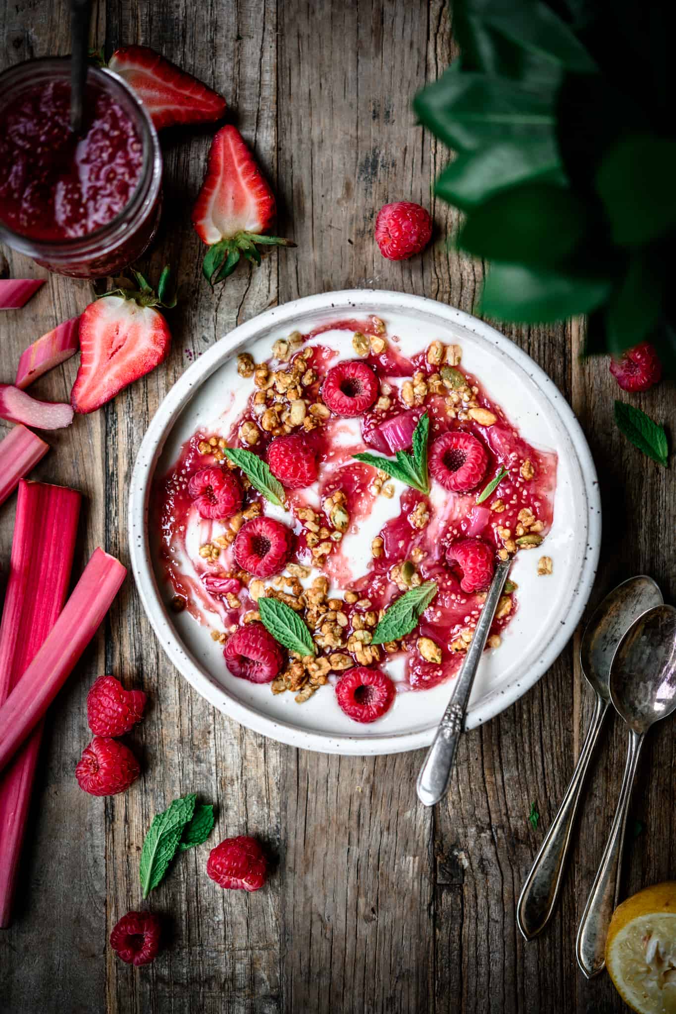 Overhead view of bowl of yogurt with rhubarb compote swirl, granola, raspberries and mint on wood table