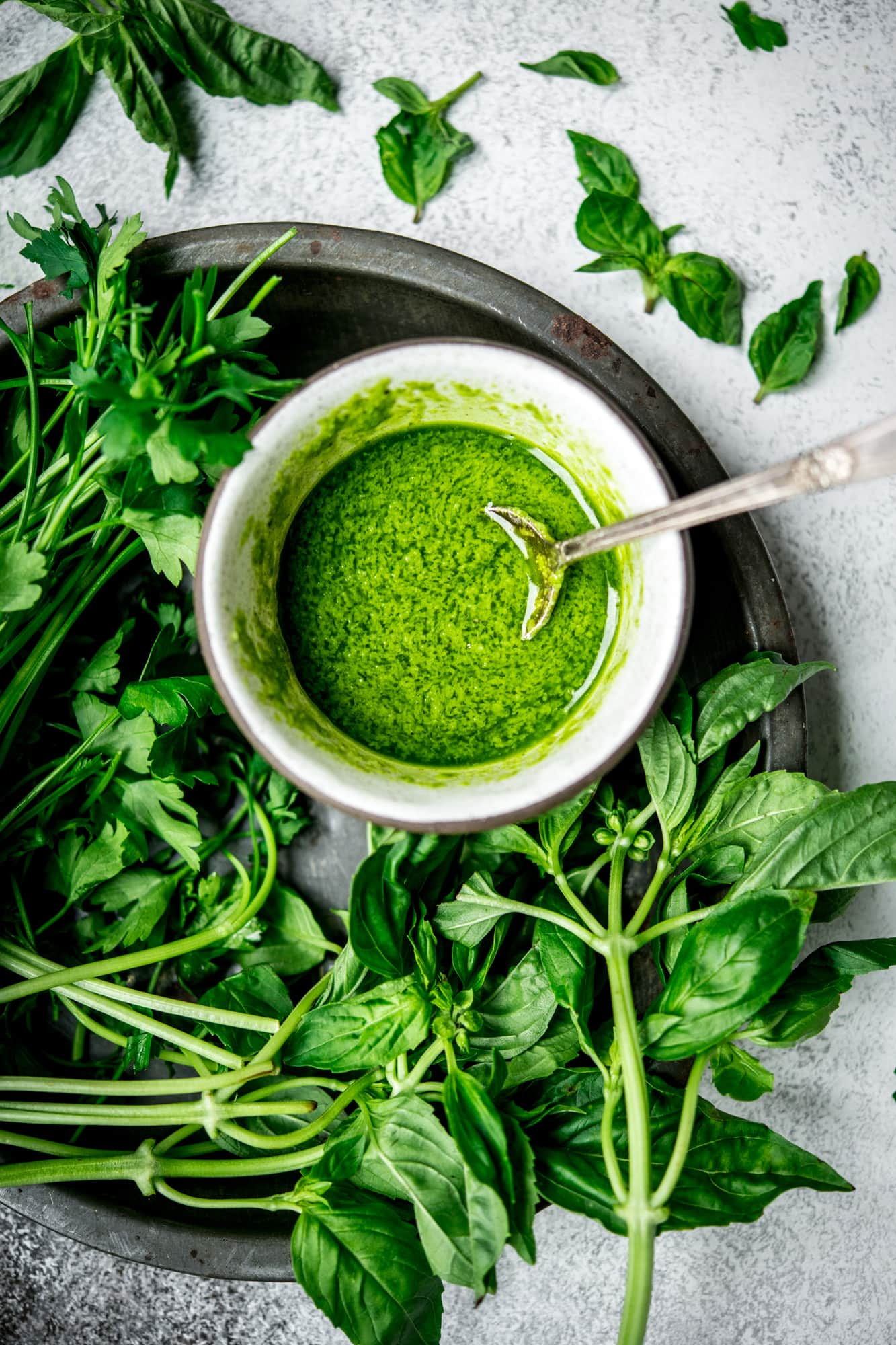 Overhead view of green herbed oil in a small white bowl surrounded by fresh herbs