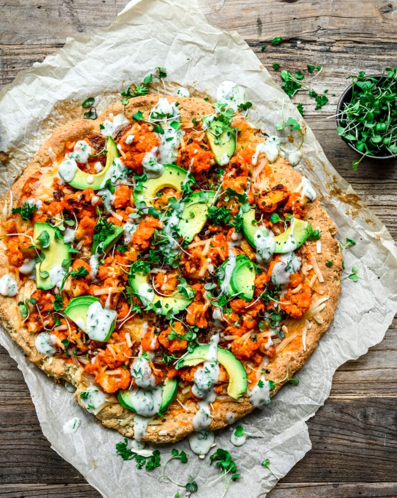 Overhead view of vegan buffalo cauliflower pizza with avocado slices and ranch drizzle on parchment paper