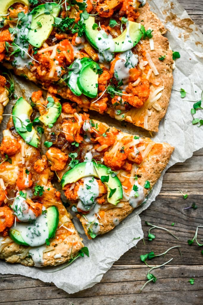 Overhead view of vegan buffalo cauliflower pizza with avocado slices and ranch drizzle on parchment paper