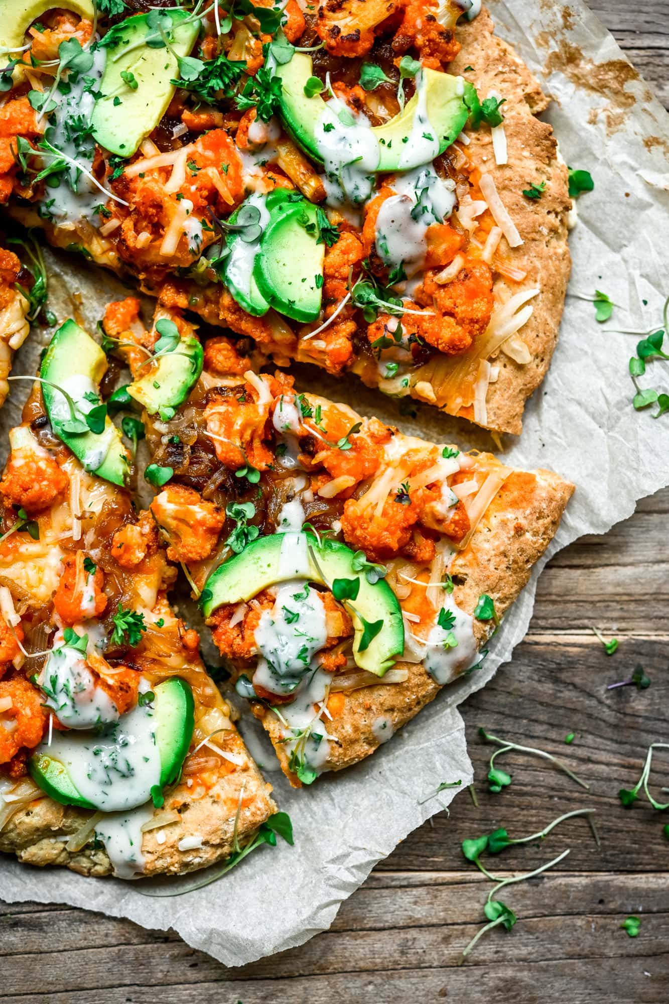 Overhead view of vegan comfort food buffalo cauliflower pizza with avocado slices and ranch drizzle on parchment paper