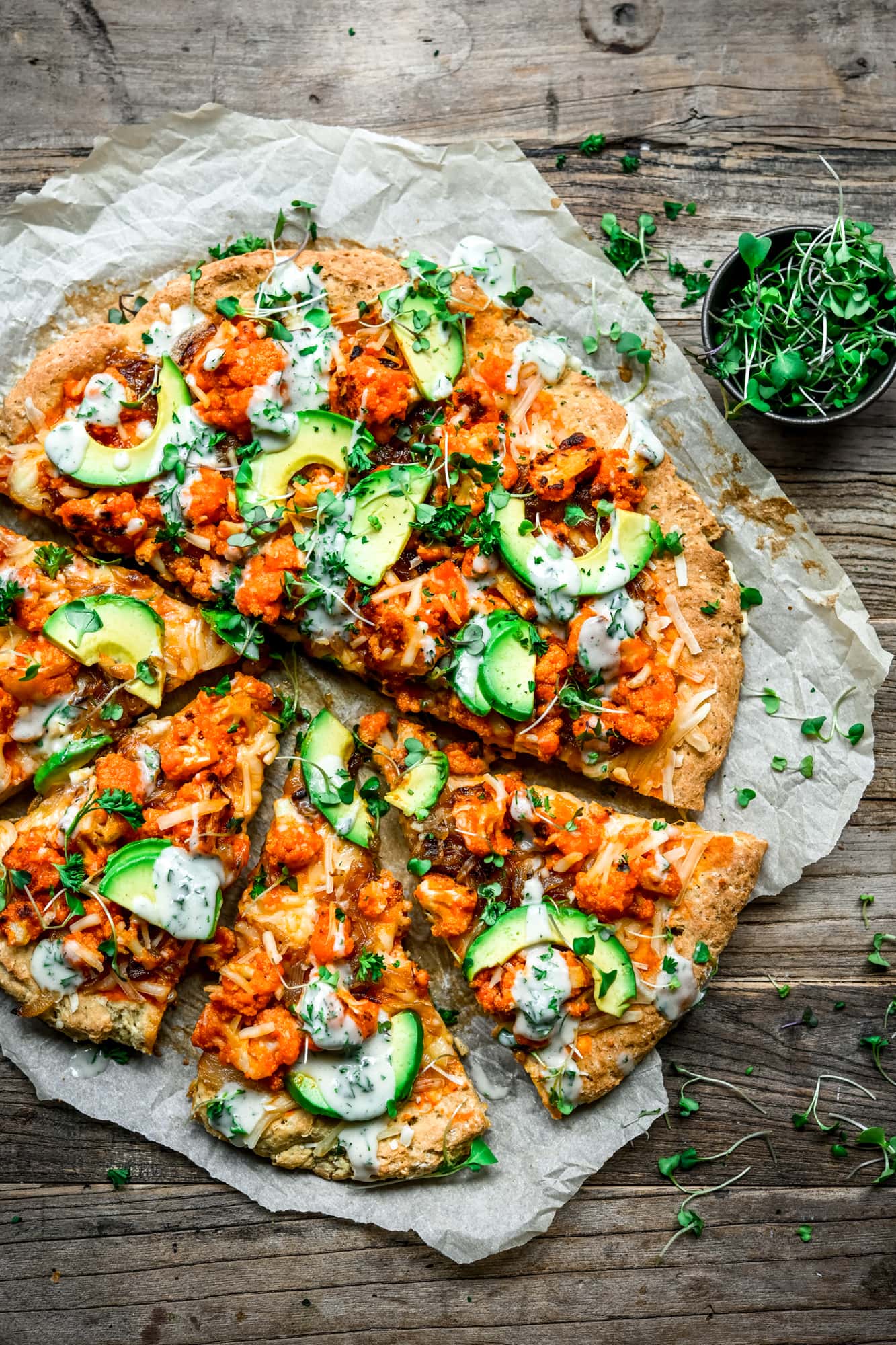 Overhead view of sliced vegan buffalo cauliflower pizza with avocado slices and ranch drizzle on parchment paper