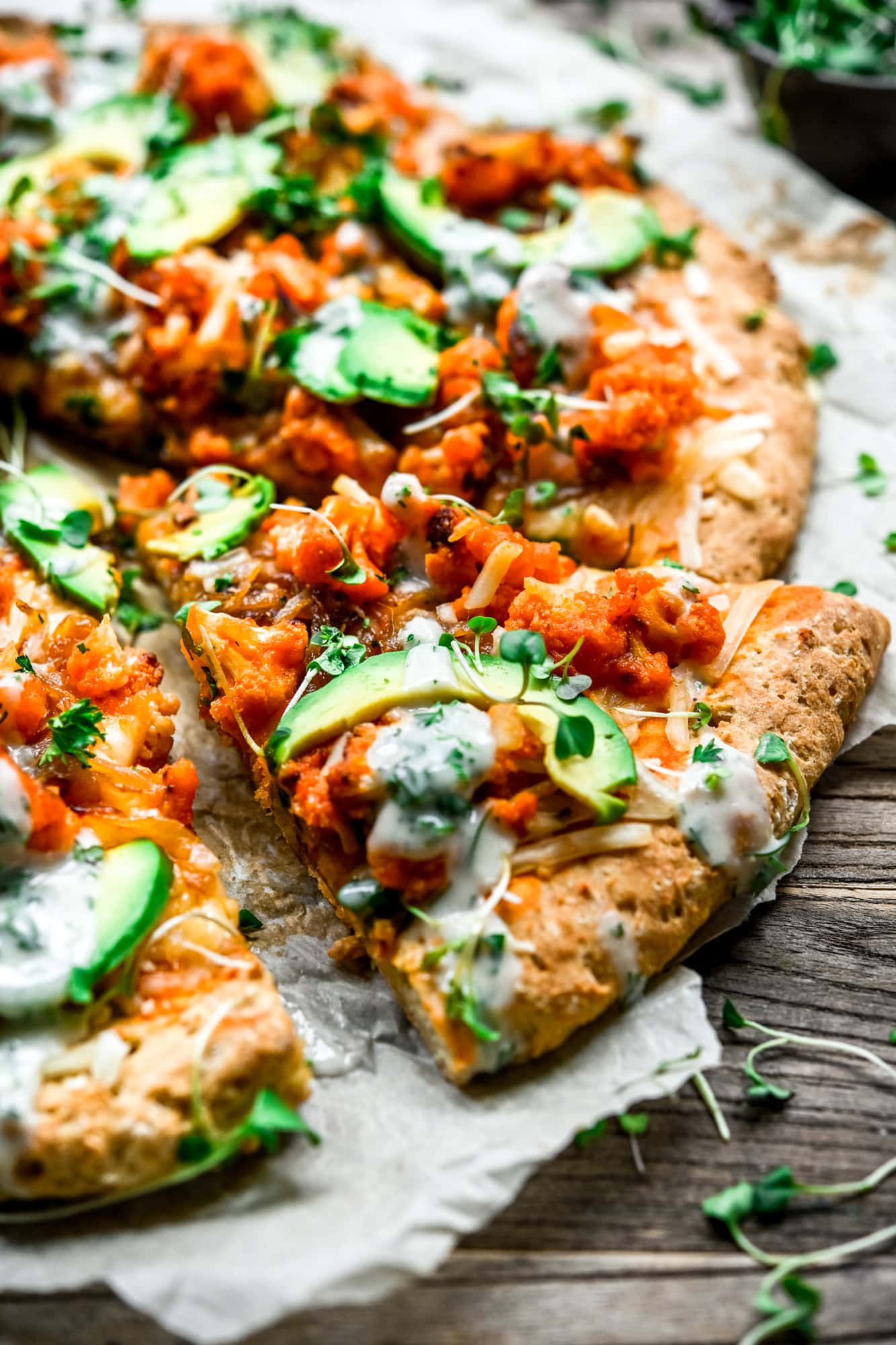 Close up side view of vegan comfort food buffalo cauliflower pizza with avocado slices and ranch drizzle on parchment paper