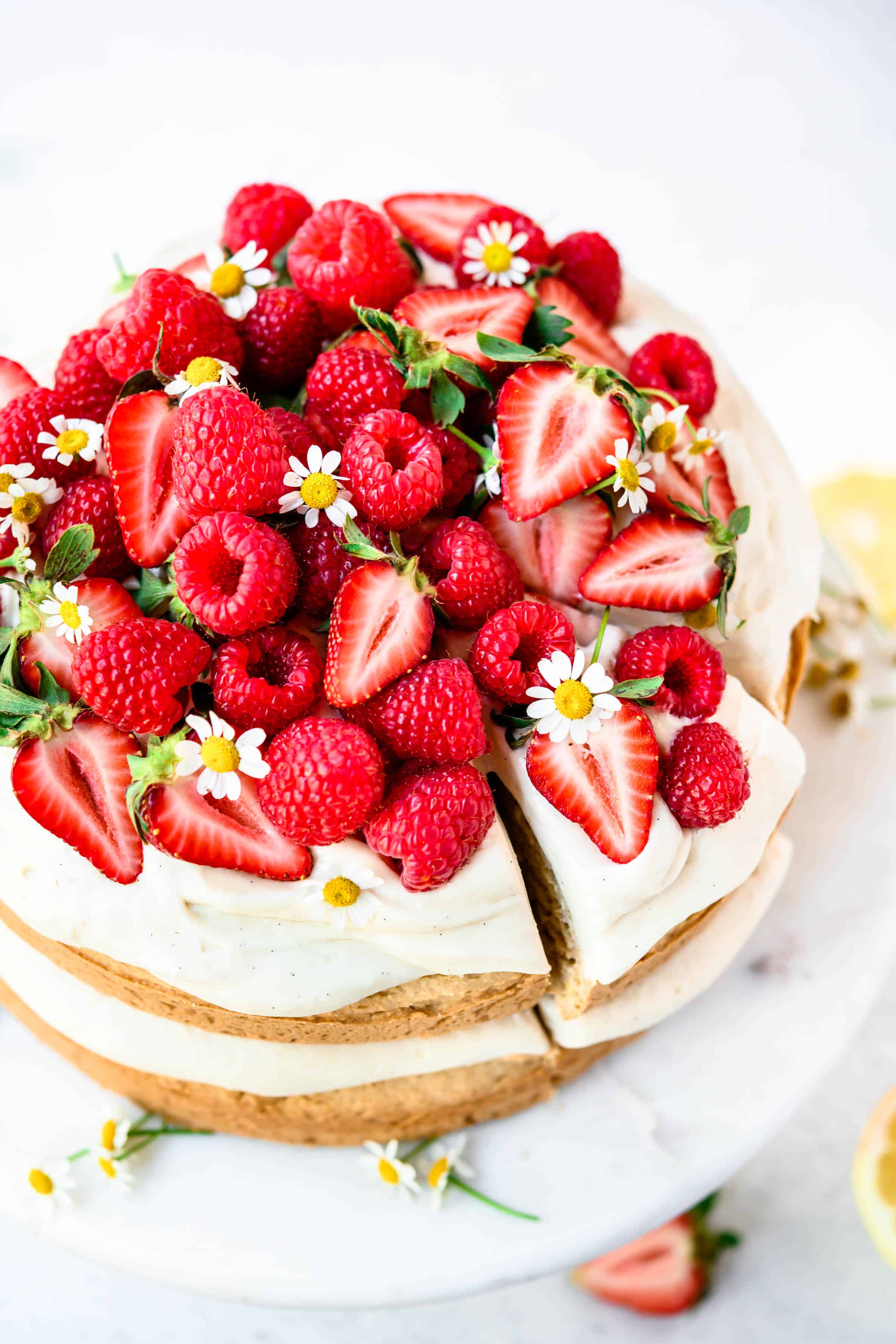 High side view of vanilla layer cake topped with fresh berries and flowers on marble cake stand