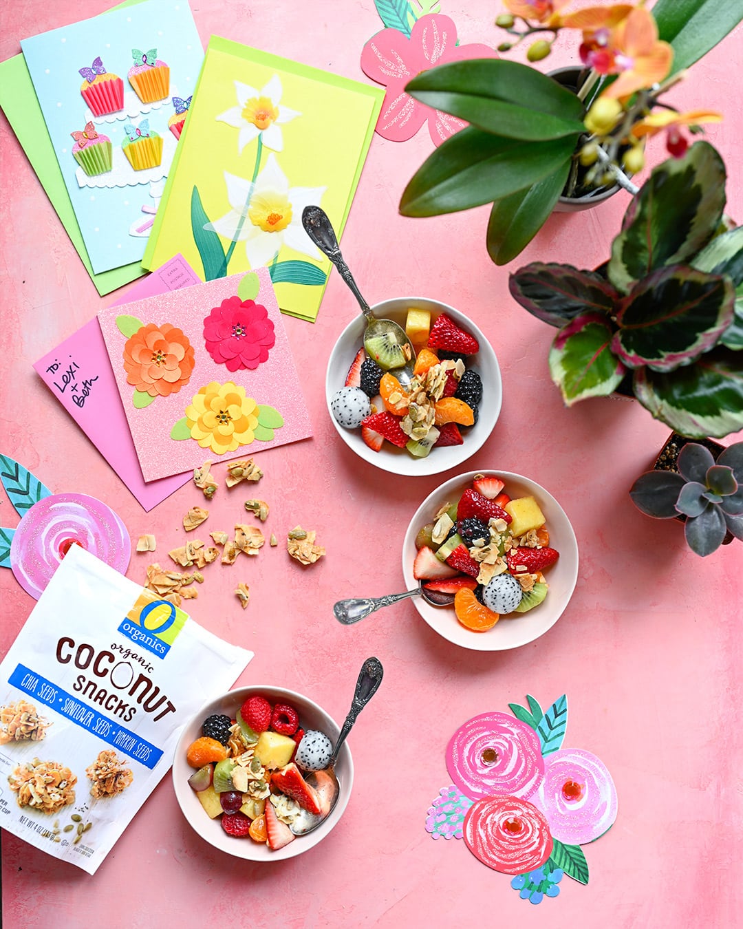 Overhead view of three fruit salad bowls on pink background with flowers and cards