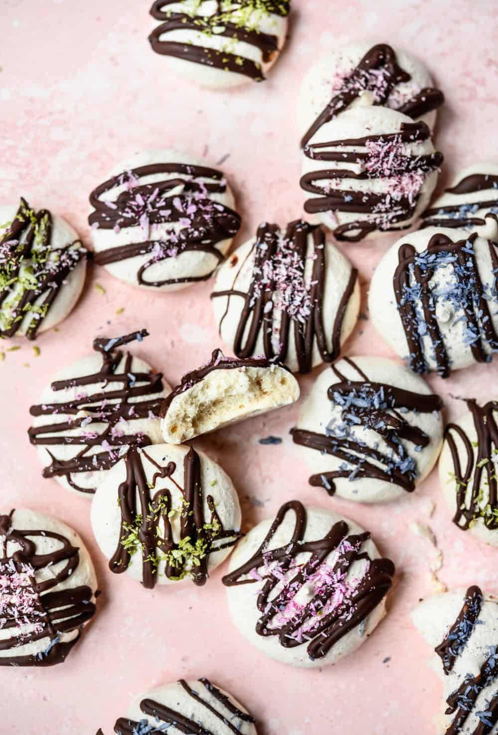Overhead view of vegan aquafaba meringues drizzled with dark chocolate and colored shredded coconut on pink background