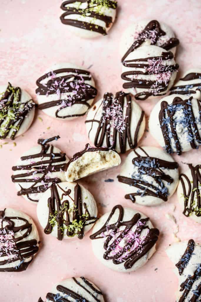 Overhead view of vegan aquafaba meringues drizzled with dark chocolate and colored shredded coconut on pink background