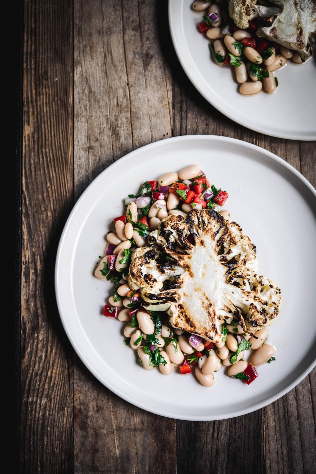 Overhead view of grilled cauliflower steaks over white bean salad on a white plate on wood table