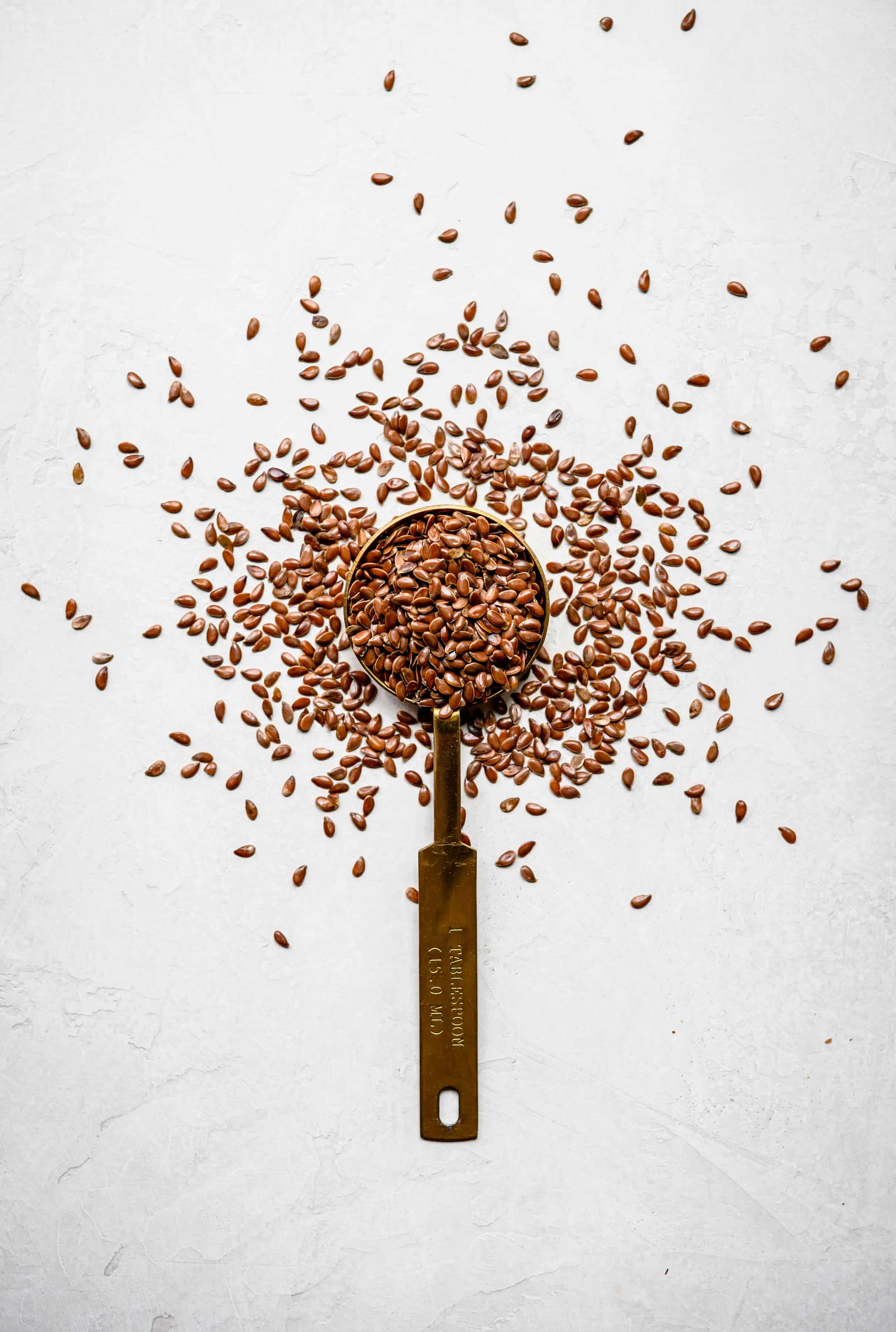 Overhead view of flax seeds on and around a gold measuring spoon on white background