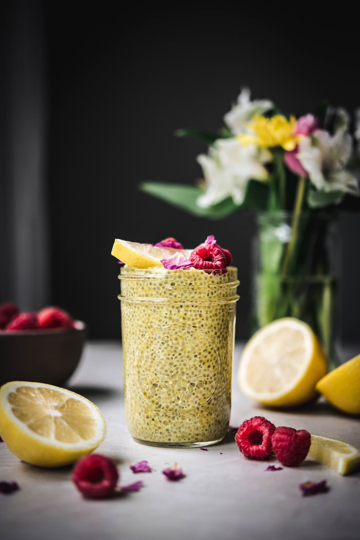 Side view of lemon turmeric chia pudding in a glass jar with fresh raspberries and flowers in background