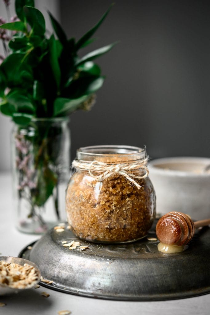 Side view of oatmeal cookie body scrub in a small glass jar with greenery in background