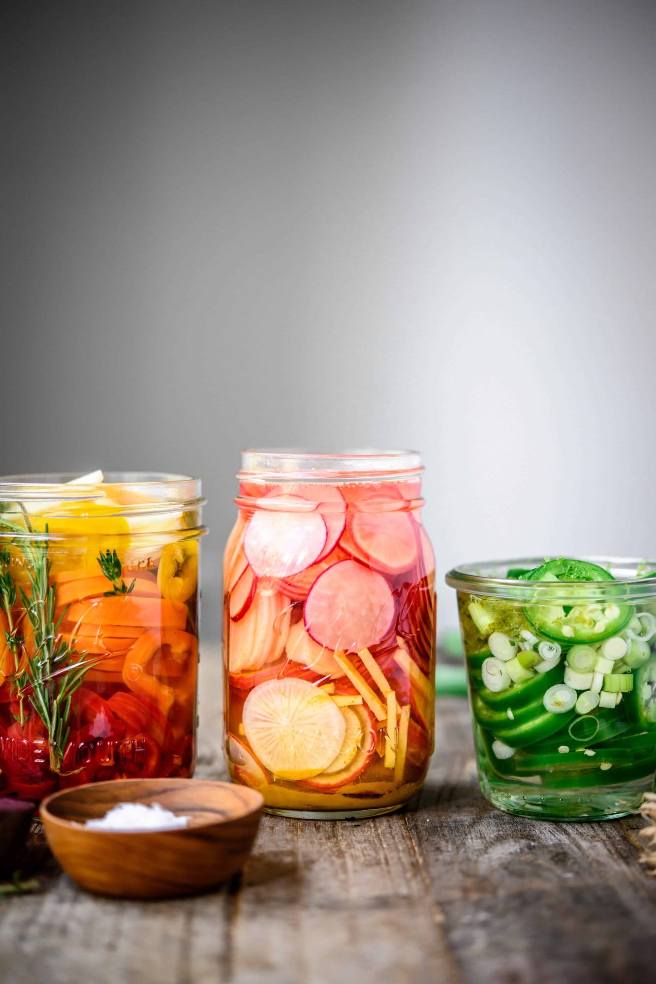 Side view of homemade quick pickled radishes, peppers and jalapeños in glass jars on wood table