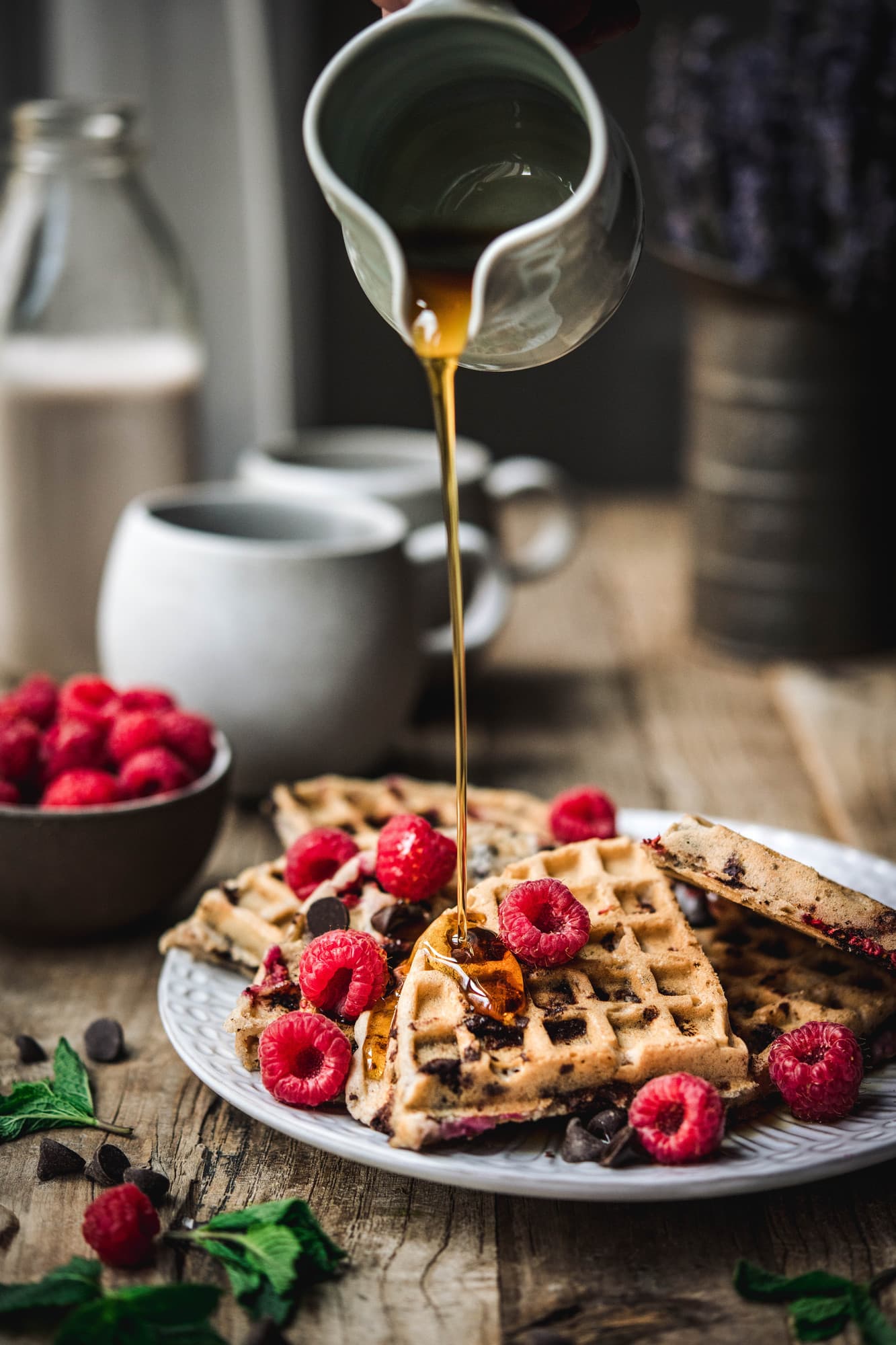 Pouring maple syrup onto raspberry chocolate chip waffles on white plate with fresh raspberries
