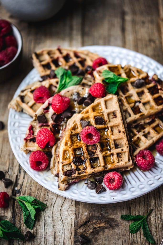 High side view of raspberry chocolate chip waffles with fresh raspberries and mint on white plate