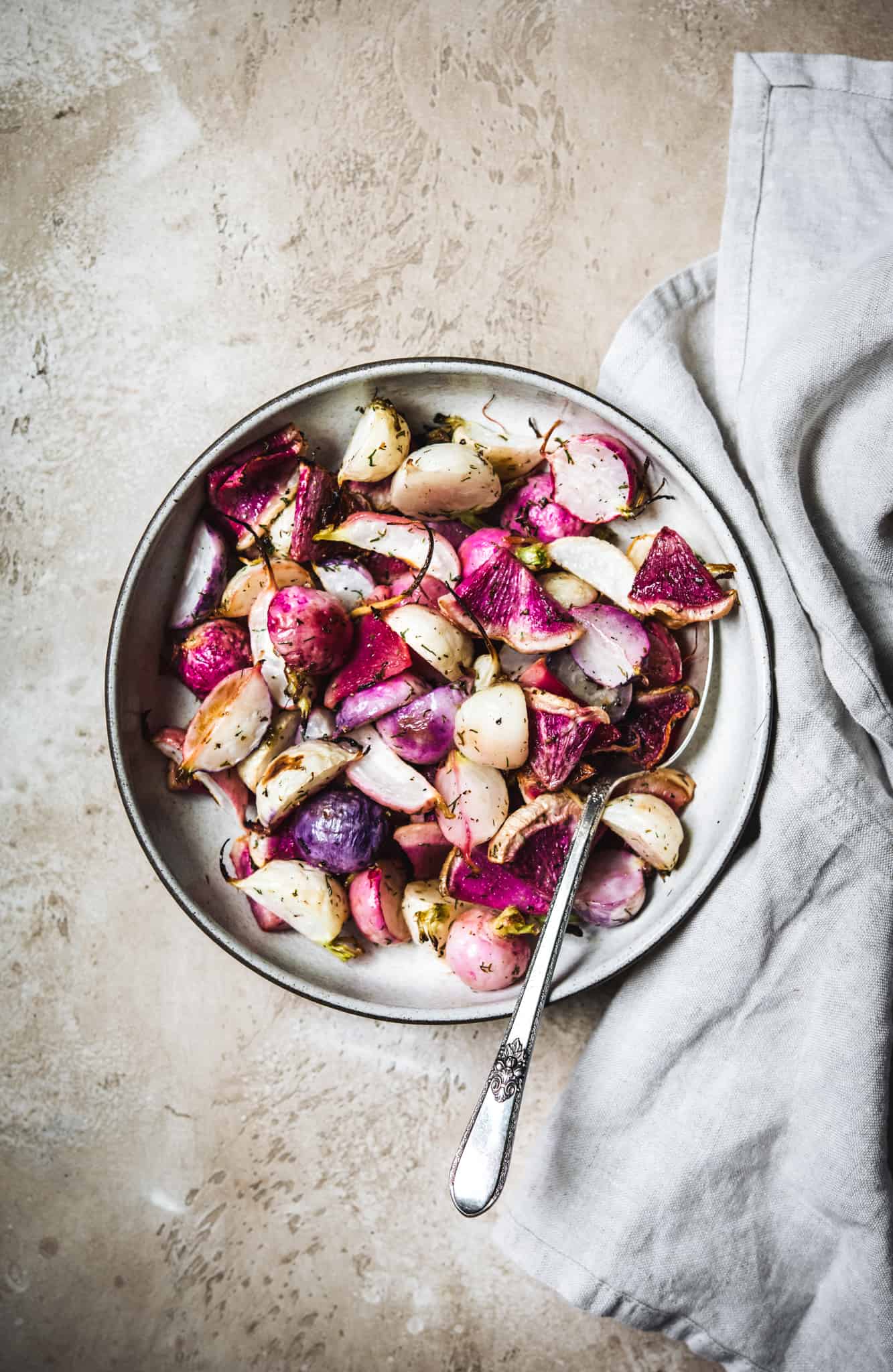 Overhead view of dill roasted radishes in a white bowl on a rustic brown background with grey towel