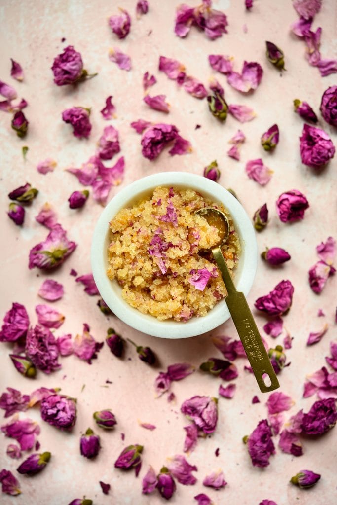 Overhead view of rose orange body scrub in white bowl on pink background with rose petals