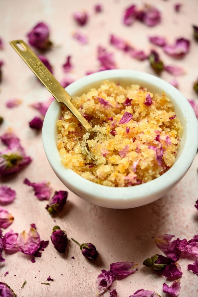 Side view of rose orange body scrub in white bowl on pink background with rose petals