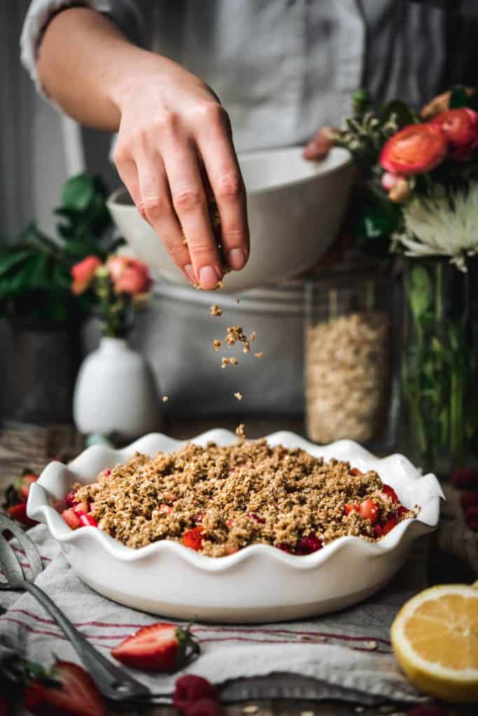 Person sprinkling crumble topping on top of strawberry rhubarb crisp with flowers in background