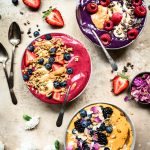 Overhead view of 3 beautiful red, purple and yellow summer smoothies bowls on rustic brown surface