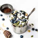 High side view of jar of maple vanilla chia pudding topped with blueberries and pepitas on white background with honey drizzler