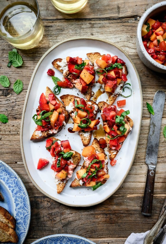 overhead view of strawberry peach bruschetta on a white ceramic platter on wood table with white wine