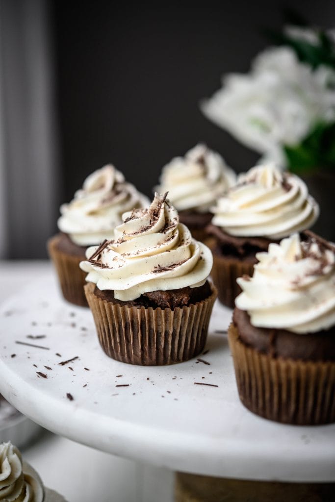 Side view of vegan chocolate cupcakes with vanilla frosting on white marble cake platter