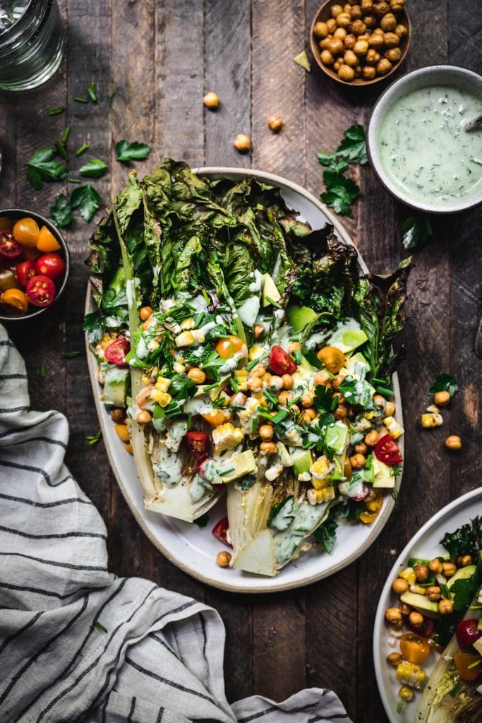 overhead of grilled romaine salad with crispy chickpeas and vegetables on an oval platter with vegan ranch dressing