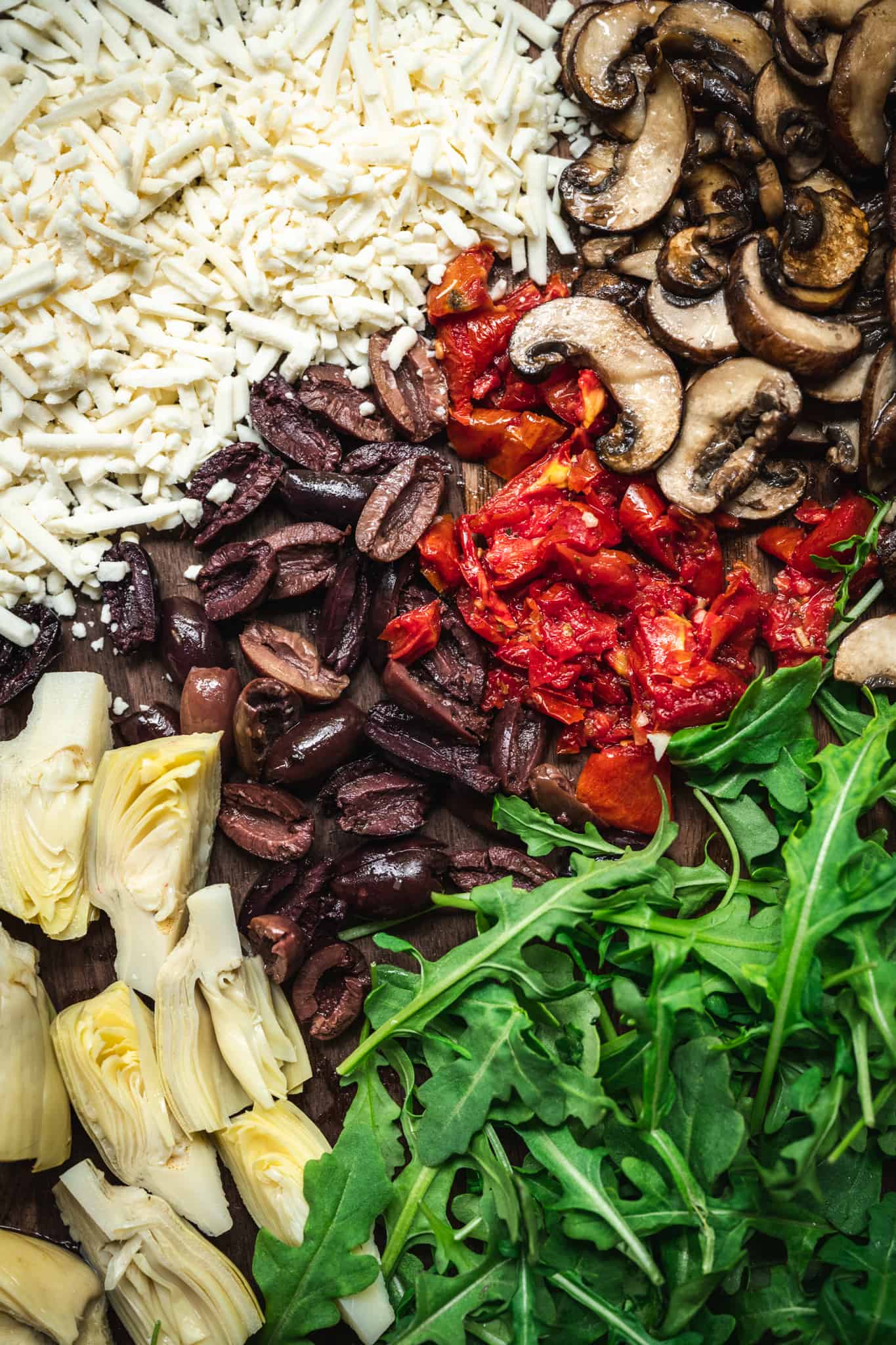 Overhead of vegan cheese, mushrooms, olives, sun-dried tomatoes, artichokes and arugula