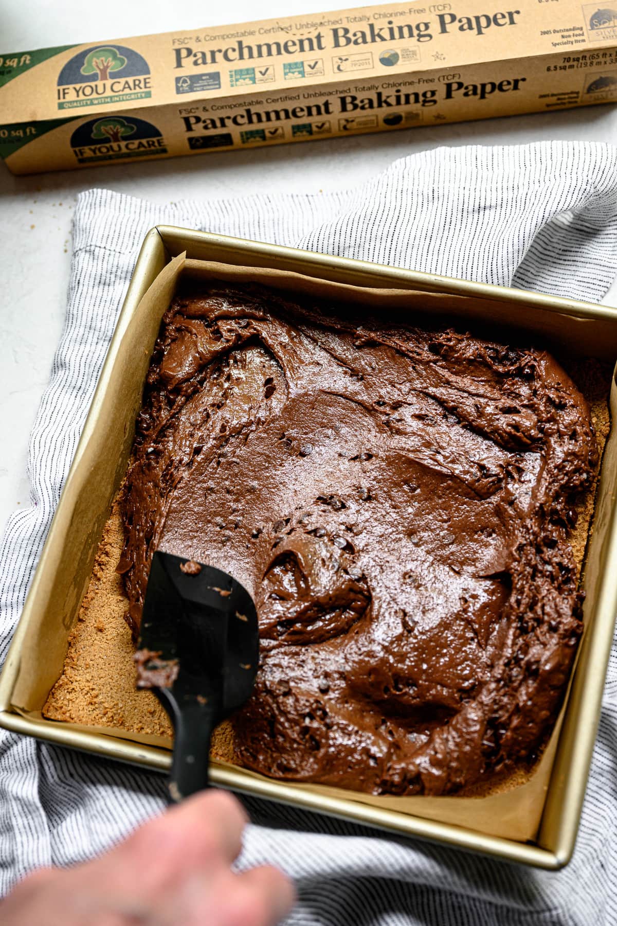 Side view of person spreading brownie batter into square pan lined with parchment paper