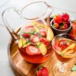 Side view of strawberry peach basil sangria in a large pitcher on wood tray
