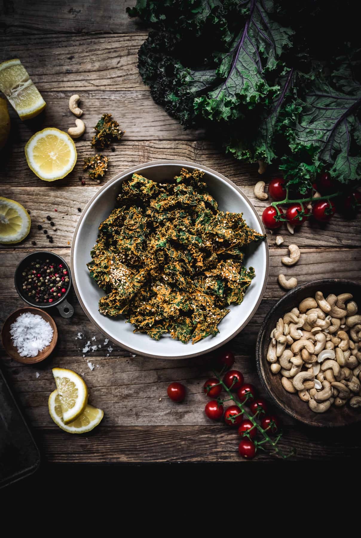 Overhead view of bowl of cheesy vegan kale chips on a wood table with ingredients around bowl