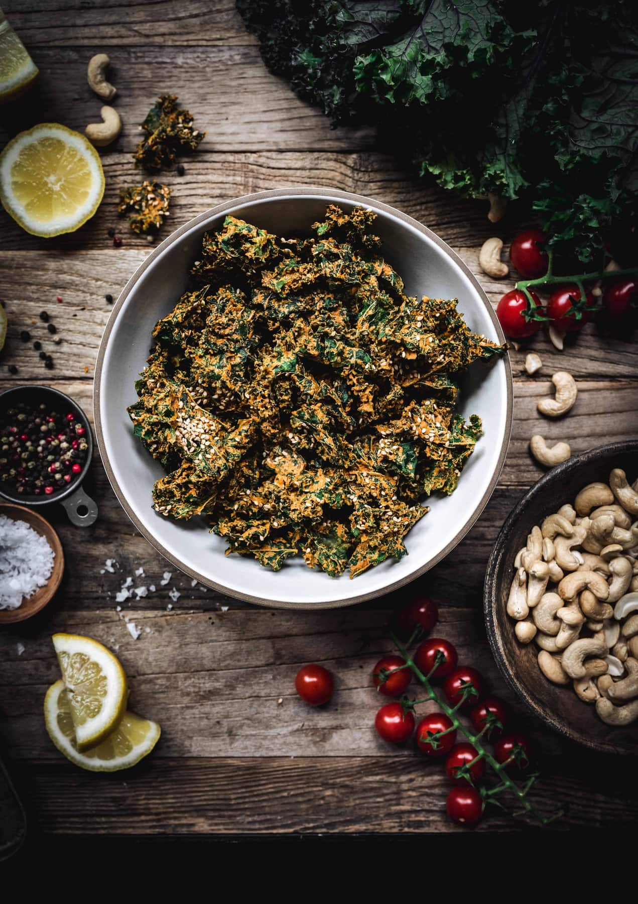 Overhead view of bowl of cheesy vegan kale chips on a wood table with ingredients around bowl