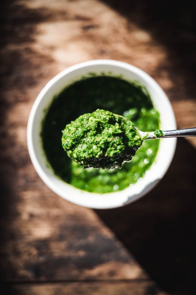 overhead of a spoonful of fresh herb pesto over a white bowl on a wooden background