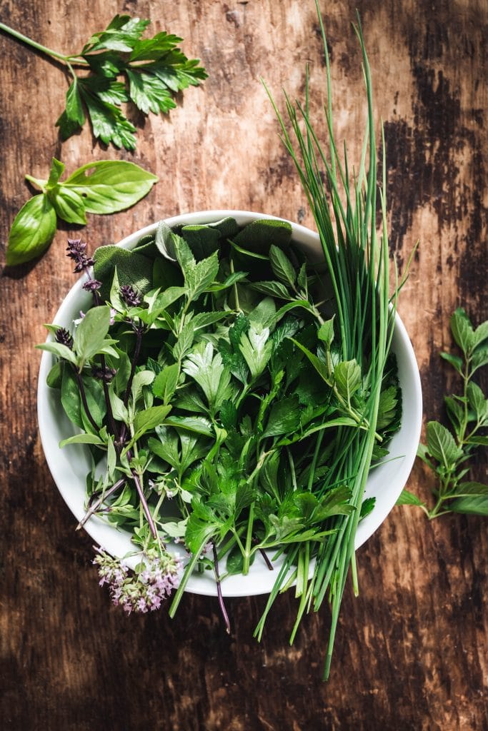 overhead of fresh herbs, oregano, basil, chives, cilantro, in a white bowl on a wooden background