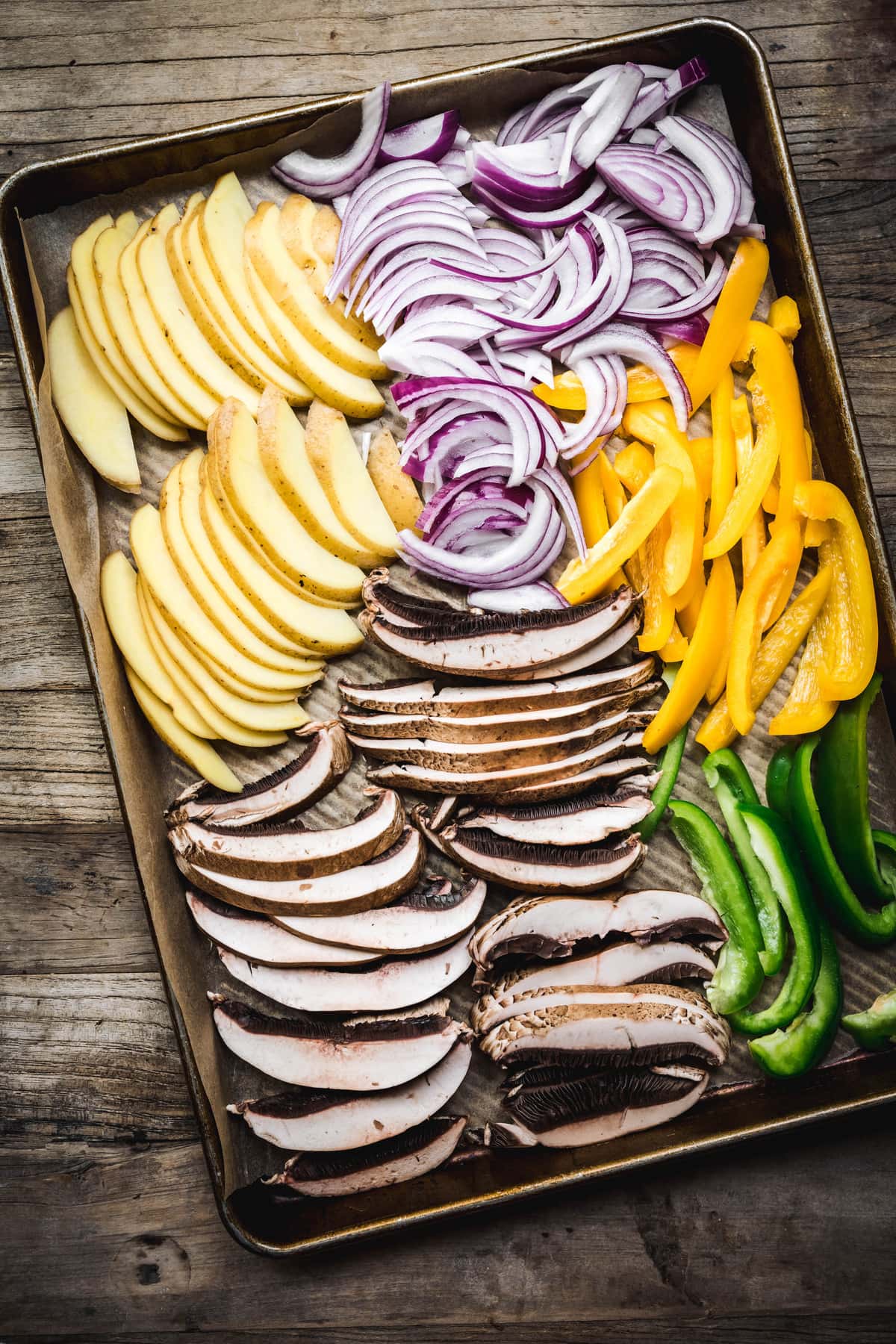 overhead of raw potatoes, portobellos, onions, and peppers on a sheet pan before cooking