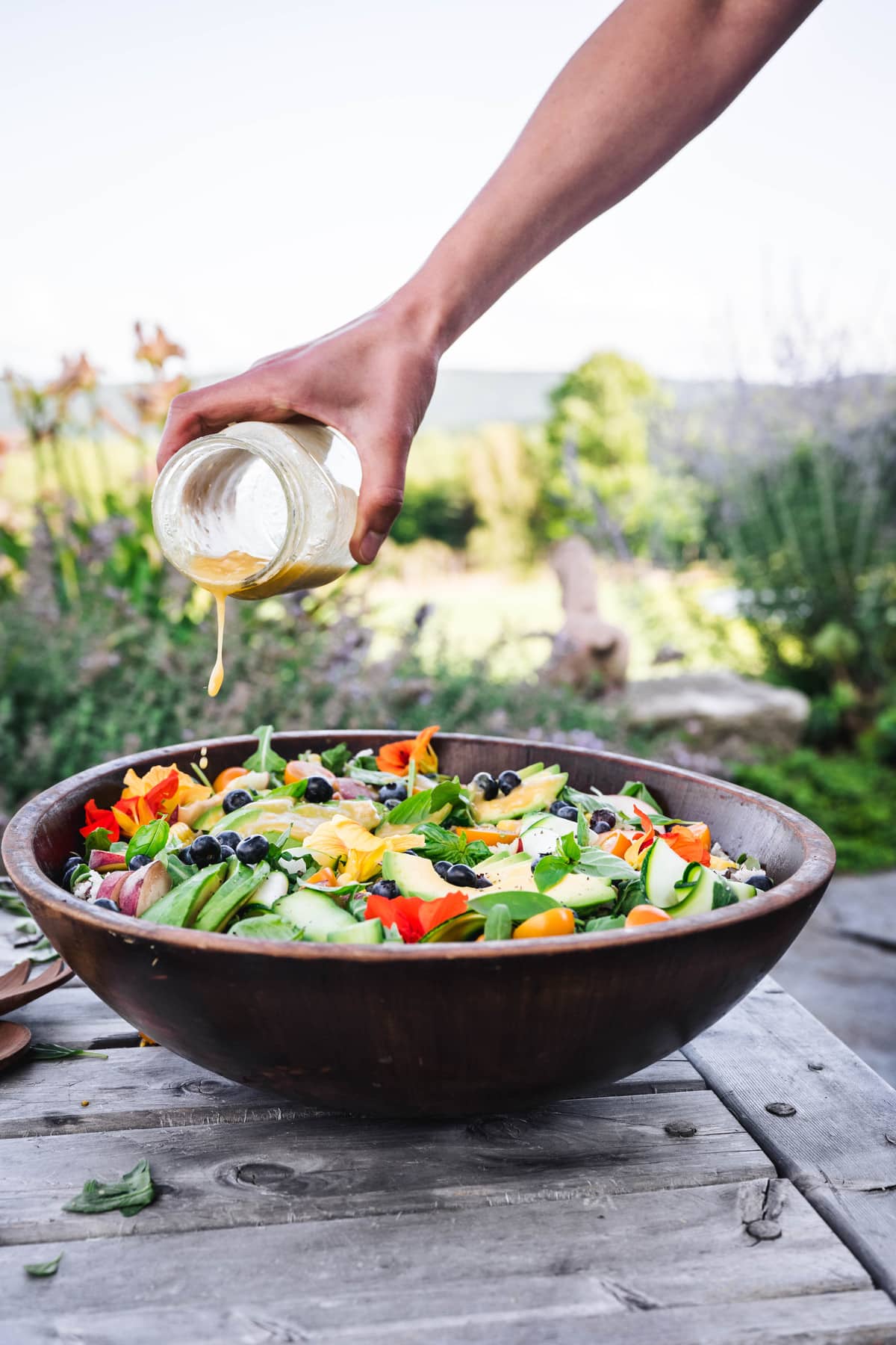 side view of someone pouring vinaigrette over summer grain salad in a wooden serving bowl with green field in the background