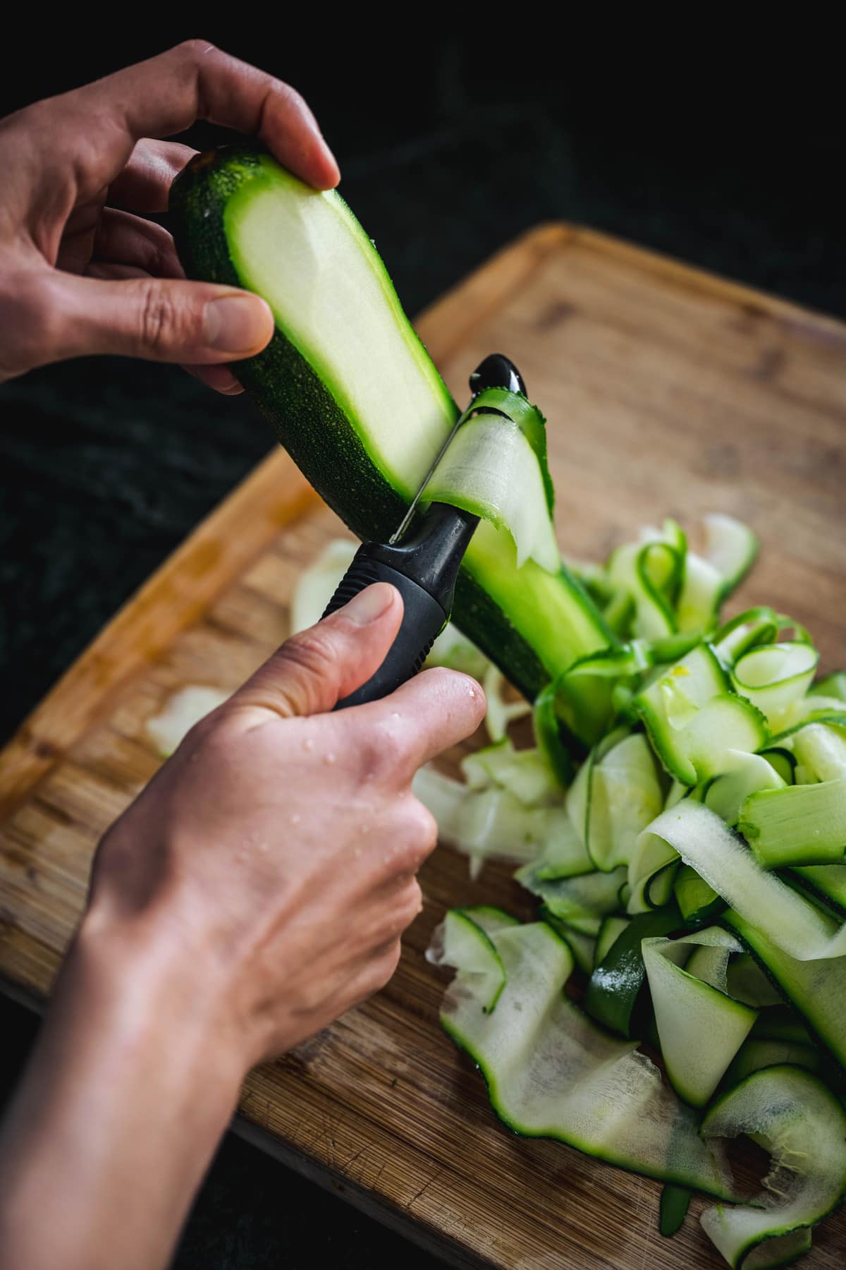 overhead of someone peeling fresh zucchini with a hand peeler on a wooden cuttingboard